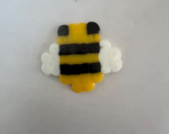 May include: A yellow, black, and white bee-shaped bead creation. The bee has a yellow body with black stripes, black head accents, and white wings. The craft item is likely made from plastic beads.