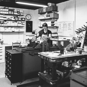 May include: A woman operates a vintage printing press in a workshop. The press is made of metal and has a large roller. The woman is wearing a black shirt and a white apron. There are shelves filled with paper and other supplies in the background.