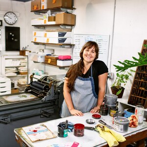 May include: A woman wearing a blue apron stands in a print shop, smiling at the camera. She is surrounded by printing equipment, including a large printing press, shelves of paper, and a variety of inks and tools.