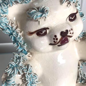 May include: A vintage ceramic poodle figurine in off-white with blue accents. The poodle has brown eyes and a dark pink mouth. The blue accents are around the ears and chest. The figurine has a glossy finish and a crackled texture.