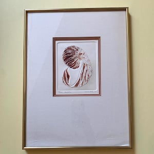 May include: A framed print featuring a copper-colored nautilus shell illustration centered on a white mat. The artwork is enclosed in a thin gold metal frame. Handwritten text and a numbering sequence are visible at the bottom of the print.