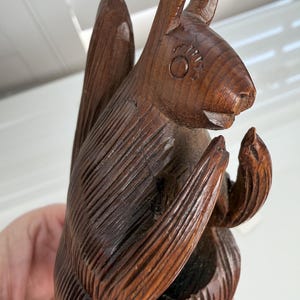 May include: A dark brown wooden figurine of a squirrel in a seated position with its front paws raised near its face. The sculpture features carved linear details representing fur texture, pointed ears, and a large, upright, curved tail.