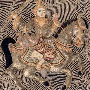 May include: An embroidered artwork depicting a figure on horseback, holding a sword. The figure and horse are adorned with sequins and intricate details. The background features swirling patterns and decorative elements, with a color palette of gold, silver, and black.