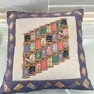 May include: Square decorative pillow with a quilted design. The center features a pattern of stacked thread spools in various colors, each with floral designs. The border is a darker blue with repeating spool patterns.