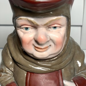 May include: A ceramic pitcher with a detailed, hand-painted design of a stern-faced man in a brown hooded robe and hat. The pitcher has a handle and a spout, and the man's face is painted with realistic features, including blue eyes and a rosy complexion.