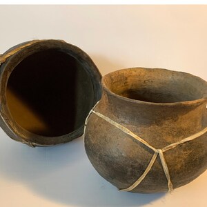 May include: Two handmade, dark brown clay pots with a rough texture. One pot is open, revealing a dark interior, while the other is upright. Both pots are secured with light-colored, woven bands.