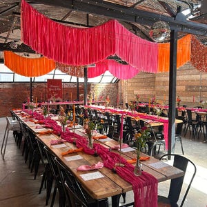 Peut inclure: Une longue table en bois dressée pour un événement spécial, ornée d'un chemin de table magenta, de fleurs et de bougies. Au-dessus, des décorations en tissu rose et orange vif sont suspendues au plafond, créant une ambiance festive. Des chaises noires sont disposées autour de la table.