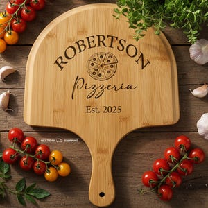 May include: A bamboo pizza peel with the text "ROBERTSON Pizzeria Est. 2025" and a pizza slice graphic. The peel is surrounded by fresh tomatoes, garlic, and herbs on a wooden surface.