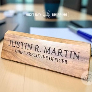 May include: A wooden desk nameplate with the engraved name "JUSTIN R. MARTIN" and the title "CHIEF EXECUTIVE OFFICER". The nameplate is rectangular and made of light-colored wood, resting on a light-colored desk surface.