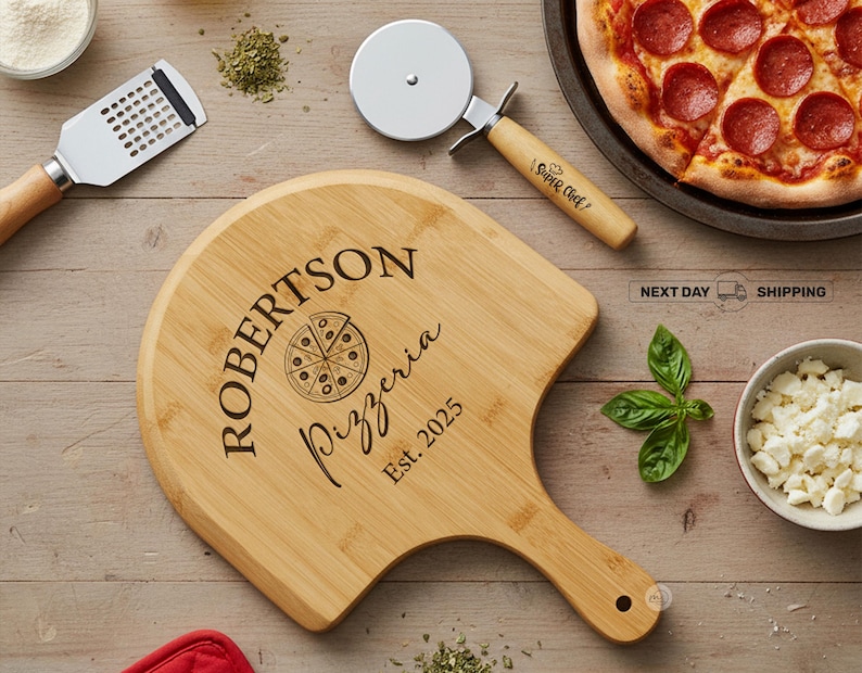 May include: A wooden pizza peel with the text "ROBERTSON Pizzeria Est. 2025" is next to a pizza cutter and a pepperoni pizza. Other items include a cheese grater, basil, and a bowl of cheese on a wooden surface.
