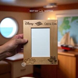 May include: A light-colored wooden picture frame with the words "Disney Cruise Line" and Mickey Mouse ears engraved on the frame. The frame has a rectangular opening for a photo and a small ship illustration.