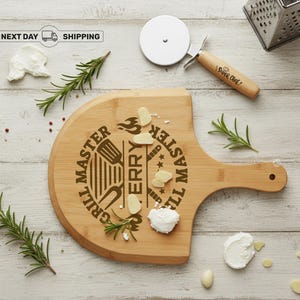 May include: A wooden pizza board with a handle, engraved with "Grill Master Terry". A pizza cutter with a wooden handle and the words "Super Chef" is next to the board. Rosemary sprigs, cheese, and garlic cloves are scattered around the board.