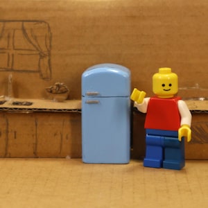 May include: A small, light blue fridge with silver handles is positioned beside a Lego figure. The Lego figure has a yellow head, a red top, white arms, blue trousers, and is waving a yellow hand. The backdrop is a cardboard kitchen scene.