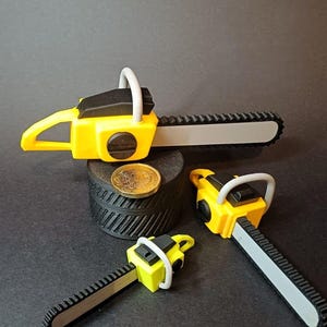 May include: A collection of miniature toy chainsaws in yellow and black, with a silver-colored blade. One is placed on a black cylindrical stand with a coin, and two others are nearby on a dark surface. The chainsaws have white handles.