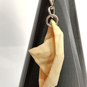 以下が含まれることがあります： 銀色のチェーンとリングに吊り下げられたベージュ色の紙のボートのキーホルダー。 紙のボートは折りたたまれ、折り目と端が見えます。 キーホルダーは、黒い幾何学的な背景に対して表示されます。