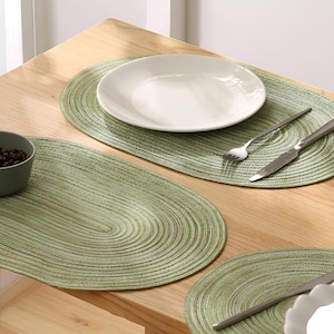 Könnte beinhalten: Ein gedeckter Tisch mit ovalen, grünen Tischsets, weißen Tellern und Besteck. Eine kleine Schale mit dunklem Inhalt ist zu sehen. Die Tischsets haben eine gewebte Textur, und der Tisch ist aus hellem Holz.