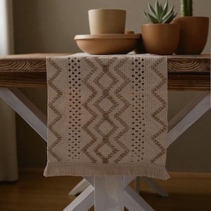 May include: A beige and brown patterned table runner with a geometric design, fringed edges, and a woven texture. The runner is displayed on a wooden table with white legs, complemented by terracotta pots and a bowl.