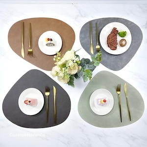 Może przedstawiać: Cztery podkładki w odcieniach brązu, szarości i zieleni, każda z białym talerzem, złotymi sztućcami i małym deserem. Bukiet białych kwiatów znajduje się pośrodku. Podkładki mają organiczny, nieregularny kształt.