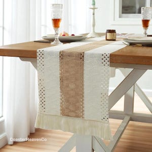 Puede incluir: Camino de mesa crema y beige con una franja central de arpillera y bordes con flecos. El camino de mesa está colocado sobre una mesa de madera, con cubiertos y cristalería visibles. El texto "CoasterHeavenCo" está en la esquina inferior izquierda.
