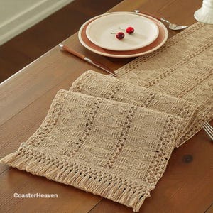 May include: A woven beige table runner with a fringed edge, placed on a wooden table. A white plate with a pink rim and two cherries sits on top. A knife and fork are placed on the table.
