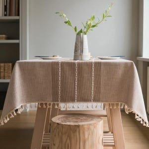 May include: A beige linen tablecloth with a tassel trim, draped over a wooden table. A vase with foliage and two plates are on the table. A tree stump stool sits beneath. The table is approximately 150 cm long.