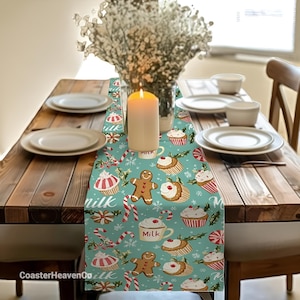 Puede incluir: Un camino de mesa navideño turquesa con un patrón festivo de hombrecitos de jengibre, cupcakes, bastones de caramelo y tazas de leche. Sobre una mesa de madera, con platos, tazas y una vela encendida.