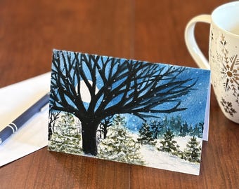 May include: A folded greeting card featuring a winter landscape painting. The artwork depicts a dark tree silhouetted against a blue night sky with a full moon. Snow-covered evergreen trees are in the foreground. A white mug with snowflake designs is in the background.