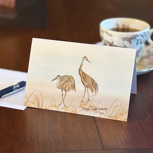 May include: Watercolor art print featuring two sandhill cranes in a natural setting. The artwork showcases the birds with detailed plumage and long legs, set against a backdrop of tall grass and a soft sky. Signed by the artist.