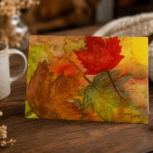 May include: A rectangular greeting card featuring a watercolor painting of autumn leaves in shades of vibrant red, golden yellow, and deep brown. The artwork displays a textured, layered effect with visible brushstrokes and a signature in the corner.