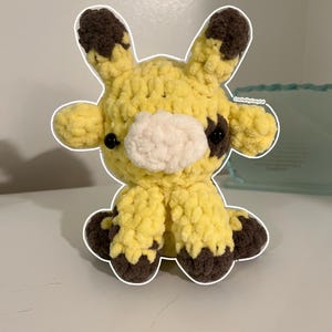 May include: A small, handmade crochet plush toy shaped like a giraffe. The body is made of soft, yellow yarn with dark brown patches on the ears, feet, and side. It features a white muzzle, two black plastic eyes, and the text CrochetbyCozyCat.