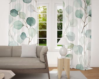 May include: White sheer curtains with a watercolor eucalyptus leaf design in shades of green and blue. The curtains hang in front of a window, with a sofa and side table in the foreground. The curtains add a touch of nature to the room.
