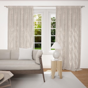 Könnte beinhalten: Beige Vorhänge mit sich wiederholendem Blättermuster hängen vor einem Fenster. Ein helles Sofa mit einem weißen Kissen steht vor den Vorhängen. Eine weiße Lampe steht auf einem kleinen Holztisch.