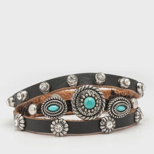 Pode incluir: Pulseiras de couro preto empilhadas com tachas prateadas e enfeites decorativos. As pulseiras apresentam pedras de cor turquesa e strass transparentes, criando um visual em camadas de inspiração ocidental.