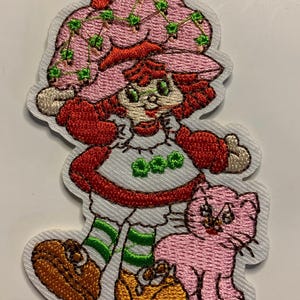 May include: An embroidered patch featuring a character with red hair wearing a large pink hat with green vine details, a white dress with green accents, green and white striped socks, and brown shoes, standing next to a small pink cat figure.