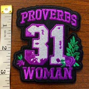 May include: A black embroidered patch featuring the purple text PROVERBS 31 WOMAN and a large silver number 31 with purple accents, decorated with green leaves and a purple flower, next to a measuring tape showing a height of 3 inches.