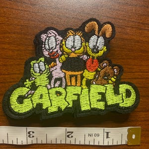 May include: An embroidered patch featuring the characters Garfield, Odie, Nermal, and Pooky above the green text GARFIELD. A white measuring tape placed below the patch shows it measures approximately 3.5 inches in total width.