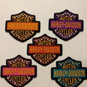 May include: Five embroidered Harley-Davidson patches in a shield shape with a checkered border. Each patch features the text MOTOR HARLEY-DAVIDSON CYCLES in different color combinations: orange, purple, red, pink, and light blue on black backgrounds.