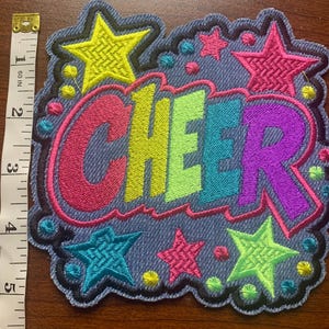 May include: A denim-textured embroidered patch featuring the word CHEER in multi-colored block letters, surrounded by neon stars and dots. A white measuring tape is placed alongside, showing the patch height is approximately 5 inches.
