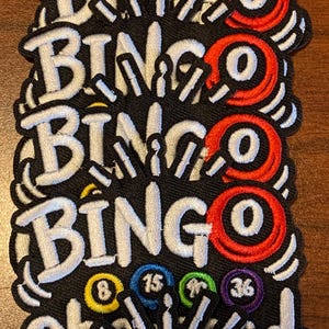 May include: A stack of embroidered patches featuring the word BINGO in white letters on a black background. Each patch includes a red bingo ball with the letter O, and smaller yellow, blue, green, and purple balls numbered 8, 15, 40, and 36.