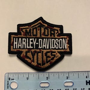 May include: A shield-shaped embroidered patch featuring the Harley-Davidson Motor Cycles logo in black, white, and a brown camouflage pattern. A ruler placed below the patch shows it measures approximately 3.5 inches in total width.