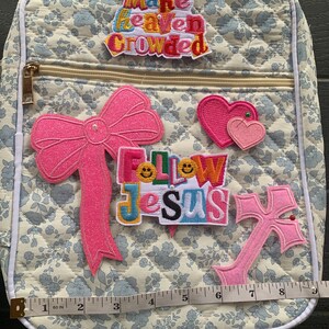 May include: A quilted backpack with a floral pattern, featuring a gold zipper and white trim. Embellishments include a pink bow, two pink hearts, and a pink cross. The text "Make Heaven Crowded" and "Follow Jesus" are also present.