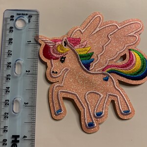 May include: A glittery peach-colored unicorn patch with wings, a rainbow mane, and a rainbow tail. The unicorn has a pink horn and blue hooves. A clear plastic ruler is positioned next to the patch, showing a height of approximately 3.5 inches.