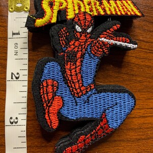 May include: An embroidered patch featuring the character Spider-Man in a dynamic pose with red and blue thread, topped with the yellow text SPIDER-MAN on a black background. A white measuring tape shows the patch is approximately 4 inches tall.