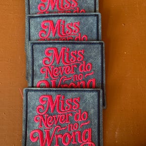 May include: A stack of denim patches, each embroidered with the red text "Miss Never do no Wrong". The patches have a dark blue border. The patches are approximately 10 cm long, as indicated by a measuring tape.