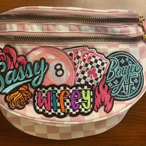 May include: A light pink and white checkered fanny pack featuring multiple embroidered patches including an eight ball, playing cards, pink flames, and roses. The patches display the text Sassy, Wifey, and Boujee AF in various colors.