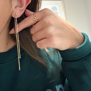 May include: Gold-toned jewelry set featuring a pair of dangling earrings and a ring. The earrings have a delicate chain design with small, round accents. The ring showcases a decorative, openwork design. The jewelry is displayed against a teal green background.