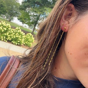 May include: Gold-colored chain earrings with a small stud at the earlobe. The earrings have a delicate, dangling chain design. The image shows the earrings on a person's ear, with long brown hair and a blue top.