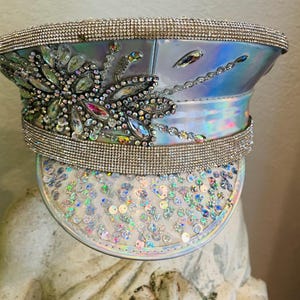 May include: Iridescent captain's hat with a sparkling rhinestone and sequin design. The hat features a large floral embellishment and a band of rhinestones around the crown. The brim is covered in sequins.
