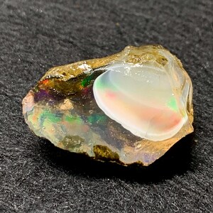 Könnte beinhalten: Ein roher Opal-Edelstein mit einem lebendigen Farbspiel. Der Opal hat eine milchig-weiße Oberfläche mit rosa, grünen und blauen Reflexen. Der Stein ist vor einem dunklen Hintergrund platziert, was seine natürliche Schönheit hervorhebt.