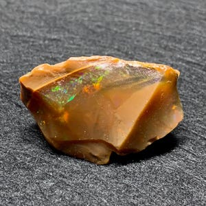 Puede incluir: Una piedra preciosa de ópalo en bruto y sin cortar, de color marrón y ámbar. El ópalo tiene destellos iridiscentes verdes y amarillos. La piedra es áspera y natural, con un aspecto facetado. El ópalo está sobre un fondo gris oscuro.